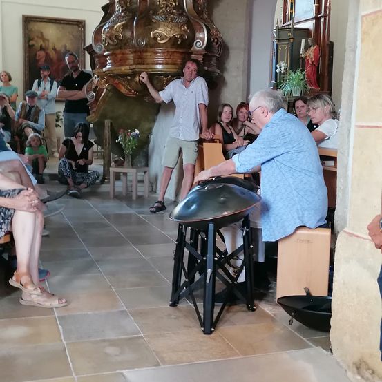 Ein älterer Mann spielt eine Gong in einer Kirche, während ein Publikum um ihn herum sitzt. Ein Mann steht hinter ihm und hält den Gong. Im Hintergrund befinden sich Gemälde und eine Statue.