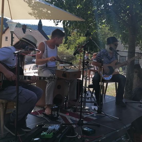 Drei Musiker spielen Gitarren auf einer Bühne unter einem Schirm, einer sitzt mit einem Fuß auf einem Lautsprecher, ein anderer steht mit Trommeln und ein Dritter steht neben einem Mikrofon in der Nähe eines Baumes.