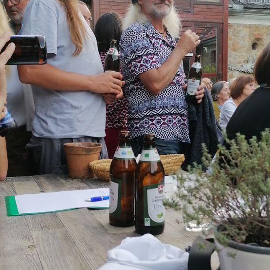 Eine Gruppe von Menschen versammelt sich um einen Tisch mit Flaschen und einer Pflanze. Eine Frau hält eine Kamera und macht ein Foto. Der Tisch hat Papiere, einen Stift und Gläser.