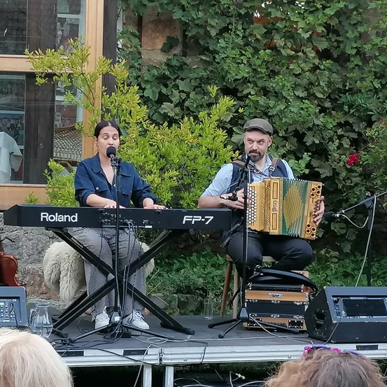 Zwei Musiker treten auf der Bühne mit Klavier und Akkordeon auf. Die Frau singt in ein Mikrofon, während sie das Klavier spielt. Der Mann sitzt auf einem Hocker und spielt das Akkordeon. Sie sind von Musikequipment und grünen Pflanzen umgeben.