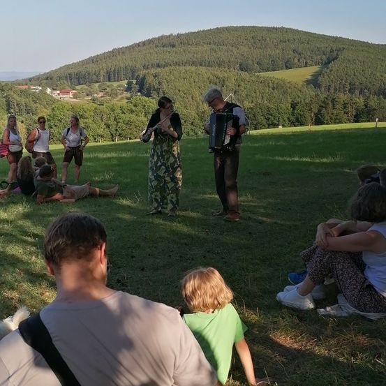 Eine Gruppe von Menschen versammelt sich auf einer Wiese, mit Bergen im Hintergrund. Zwei Menschen spielen Musikinstrumente, eine Flöte und ein Akkordeon, während andere sitzen oder stehen. Einige tragen Brillen, Hüte und Turnschuhe.