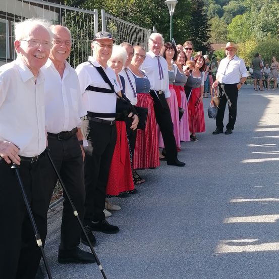 Eine Gruppe älterer Menschen in traditioneller Kleidung steht in einer Reihe. Sie lächeln und posieren für ein Foto. Sie befinden sich auf einem Betonweg mit einem Metallzaun und Bäumen im Hintergrund.
