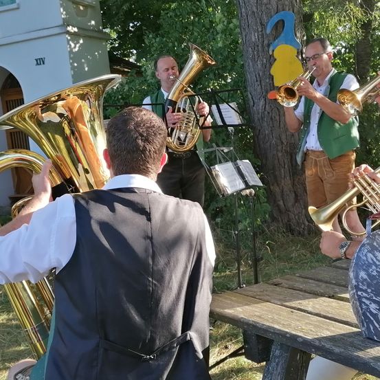 Vier Musiker spielen im Freien in einem Garten Blechblasinstrumente. Hinter ihnen ist ein Baum mit einem Nummer-2-Schild und ein Gebäude zu sehen.