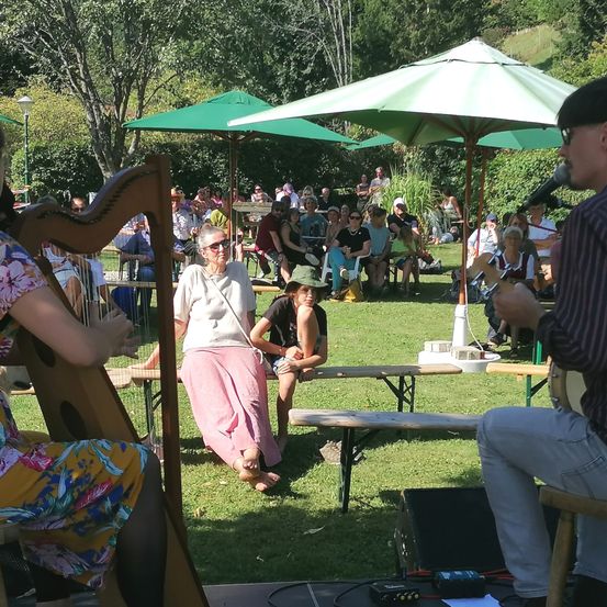 Eine Frau spielt Harfe und ein Mann Gitarre. Sie stehen auf einer Bühne in einem Park mit einem Publikum im Hintergrund. Der Park ist von Bäumen umgeben.