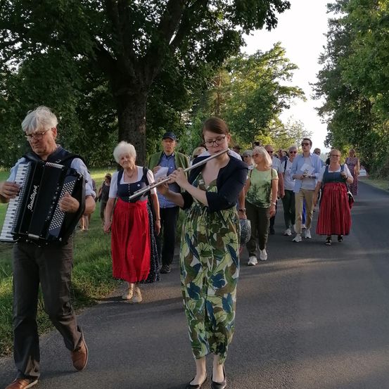 Eine Gruppe von Menschen geht auf einer Straße, wobei ein Mann ein Akkordeon spielt und eine Frau eine Flöte spielt.