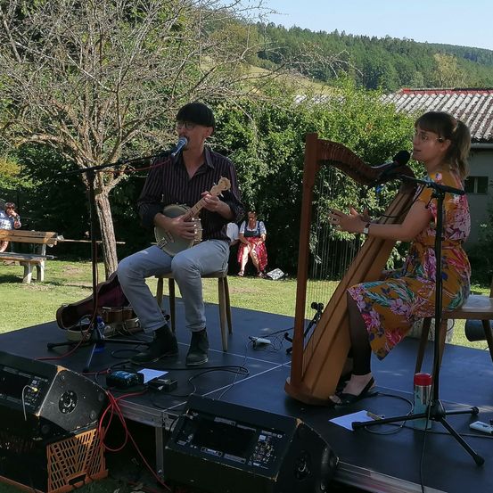 Ein Mann spielt Banjo und singt, während eine Frau eine Harfe auf einer Bühne mit grünem Hintergrund spielt.