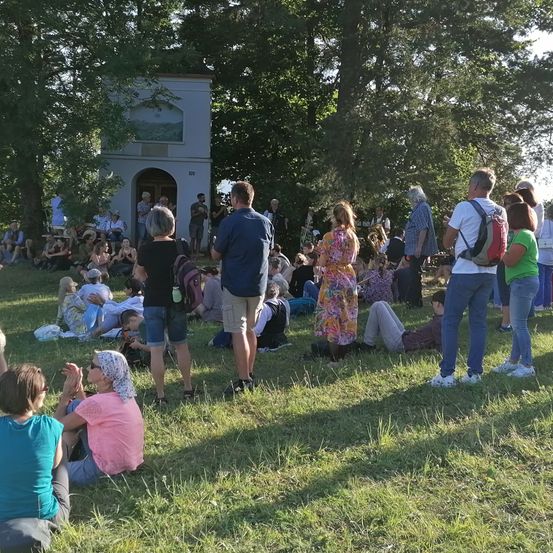 Auf einem Grasfeld im Freien sitzen oder stehen mehrere Menschen. Ein kleines Gebäude mit Tür und Fenster befindet sich im Hintergrund. Ein paar Leute stehen in der Nähe des Gebäudes. Einige Bäume umgeben das Gebiet.