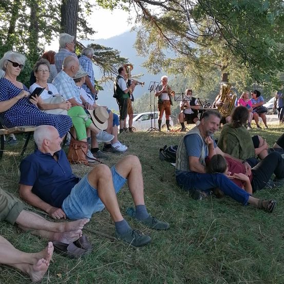 Eine Gruppe von Menschen sitzt auf dem Gras in einem Park und beobachtet eine Brassband, die spielt. Die Band steht unter einem Baum und das Publikum sitzt vor ihnen. Ein Auto ist in der Nähe geparkt.