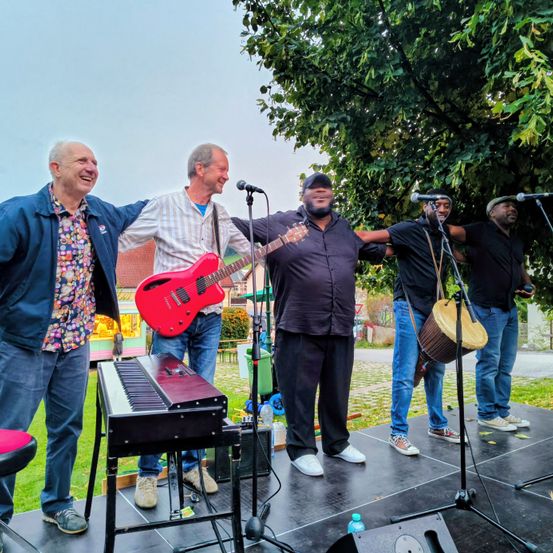 Fünf Männer spielen auf der Bühne mit Musikinstrumenten. Ein Mann spielt die Gitarre, während ein anderer ein Schlagzeug spielt. Sie haben Mikrofone vor sich. Eine Tastatur ist auf der linken Seite der Bühne.