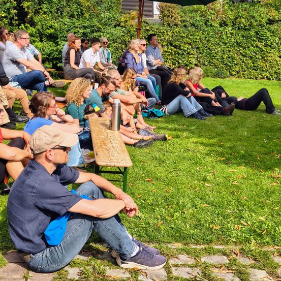Eine Gruppe von Menschen sitzt auf dem Rasen, einige auf Bänken. Einige von ihnen tragen Hüte und Sonnenbrillen. Eine Person vorne trägt eine Baseball-Kappe und Turnschuhe. Ein Holztisch mit einem Edelstahl-Becher steht auf dem Rasen. Darüber befindet sich ein grüner Schirm.