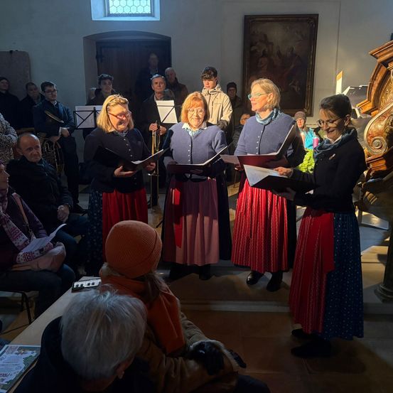 Eine Gruppe von Frauen in traditioneller Kleidung singt und hält Bücher in der Hand in einem Raum. Hinter ihnen spielt ein Mann Trompete. Der Raum hat eine Holztür, ein Gemälde und eine Holzstruktur. Mehrere Menschen beobachten sie.