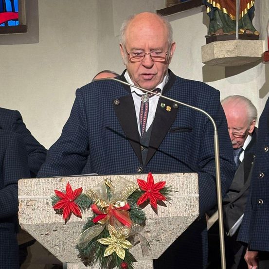 Ein älterer Mann in einem karierten Anzug steht an einem mit roten Blumen geschmückten Rednerpult und hält eine Rede. Dahinter ist eine Statue und ein Buntglasfenster zu sehen. Zwei Männer in Anzügen stehen in der Nähe.