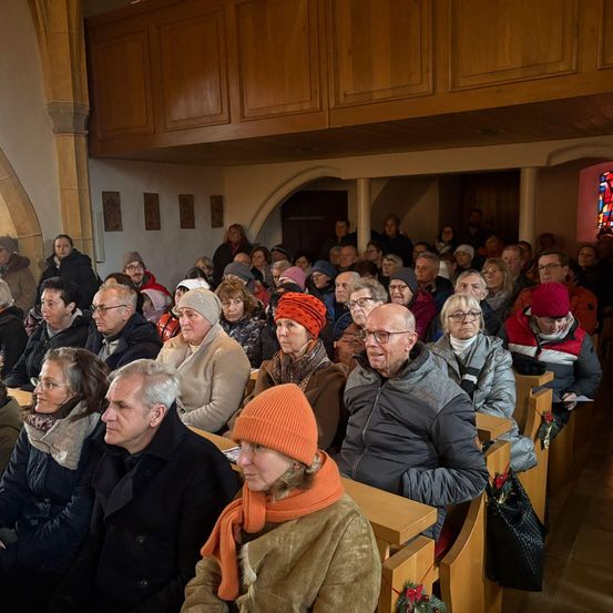 Eine große Menschenmenge älterer Menschen versammelt sich in einer Kirche. Sie sitzen in Reihen und tragen Winterkleidung. Die Kirchenbänke sind aus Holz und die Wände mit Kunstwerken geschmückt. Ein Buntglasfenster ist auf der rechten Seite zu sehen.