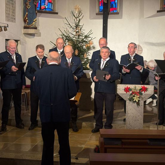 Eine Gruppe von Männern in Anzügen und Krawatten steht in einer Kirche und singt, während sie Bücher halten. Sie stehen vor einem Weihnachtsbaum und einem mit Blumen geschmückten Podium.