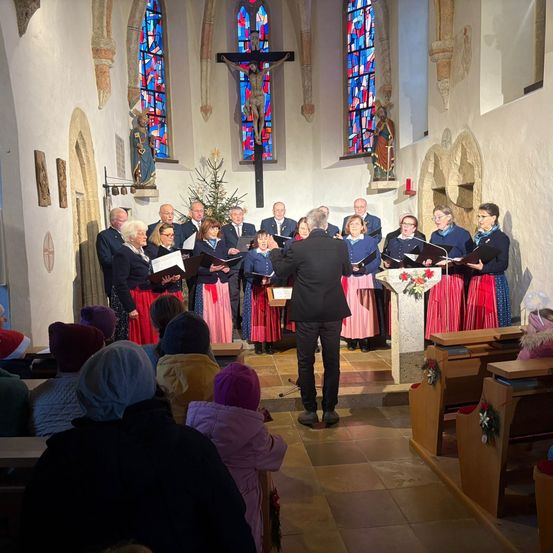 Ein Chor singt in einer Kirche mit Buntglasfenstern, angeführt von einem Mann in einem schwarzen Anzug. Die Mitglieder tragen traditionelle Kleidung, und der Altar ist mit Weihnachtsdekoration geschmückt.