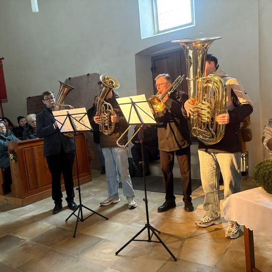 Vier Männer spielen Blechblasinstrumente in einer Kirche. Sie stehen vor Notenständern, einer der Männer trägt eine Brille. Das Publikum sitzt im Hintergrund.