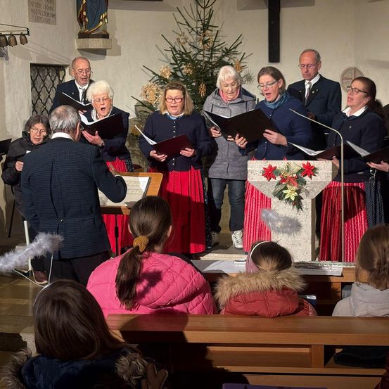Ein Chor von Menschen in Winterkleidung singt in einer Kirche. Ein Podest mit einem Weihnachtsbaum und Blumen steht vor ihnen. Menschen sitzen auf hölzernen Bänken im Vordergrund.