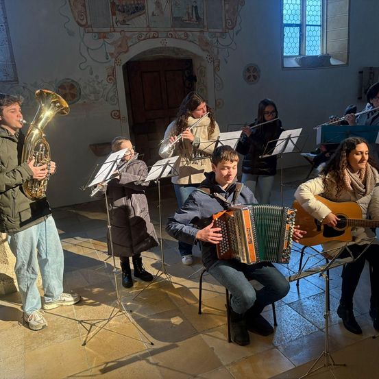 Eine Gruppe von Musikern spielt Instrumente in einer Kapelle. Ein Mann spielt Tuba, während er auf einem Hocker sitzt. Ein anderer spielt Akkordeon, während ein weiterer eine Gitarre spielt.