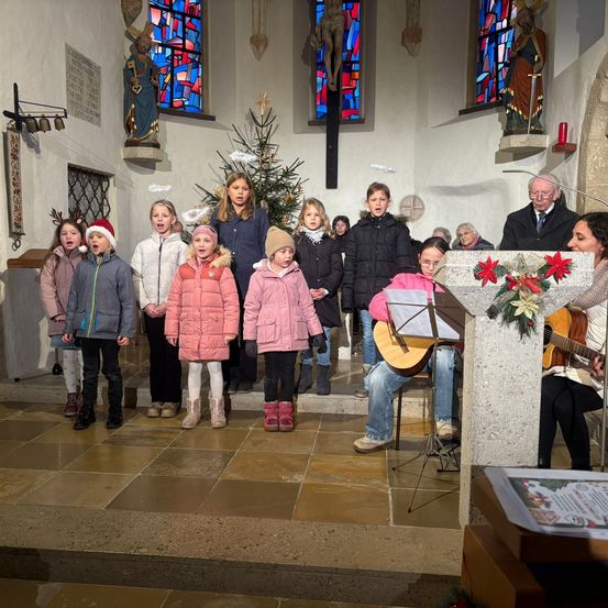 Eine Gruppe von Kindern in Winterkleidung steht in einer Kirche und singt ein Weihnachtslied, begleitet von einem Mann, der Gitarre spielt, und einer älteren Frau, die anführt. Hinter ihnen schmücken Buntglasfenster und Statuen die Wände.