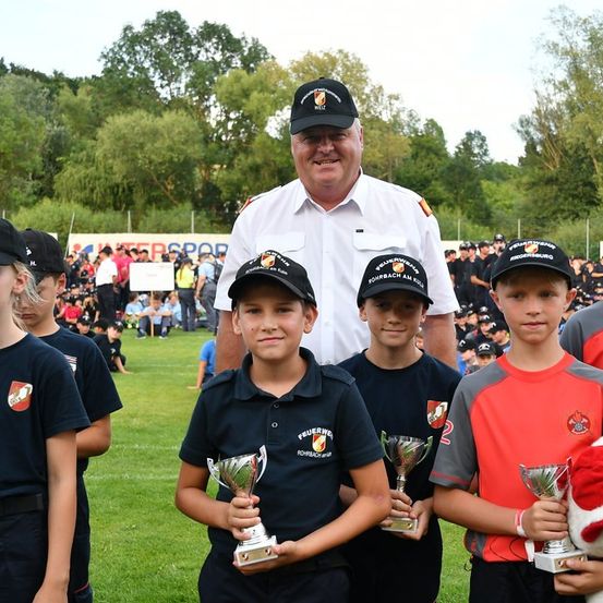 Eine Gruppe von Kindern, alle mit Mützen, stehen in einem Grasfeld und halten Trophäen. Ein Mann in einem weißen Uniform steht hinter ihnen. Bäume und ein Banner sind im Hintergrund.