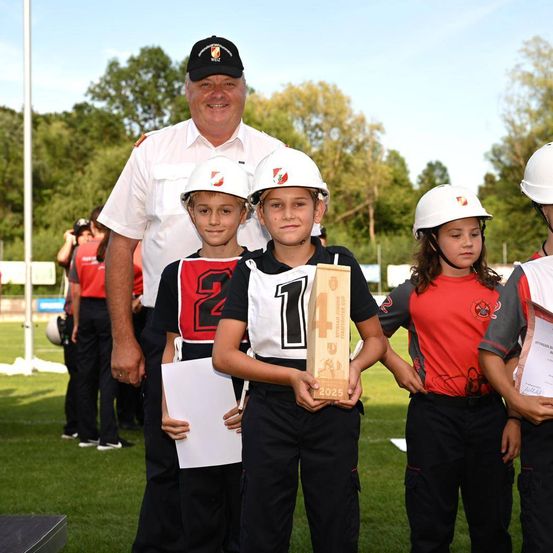 Vier Kinder in Helmen stehen in einer Reihe mit einem Mann in Uniform. Die Kinder halten Urkunden und der Mann ein Siegerpokal. Sie sind auf einem grünen Feld mit Bäumen im Hintergrund.