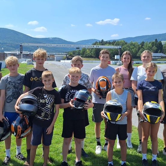 Eine Gruppe junger Menschen, sowohl Jungen als auch Mädchen, lächeln und posieren für ein Foto in einem Grasbereich, während sie Helme halten. Dahinter befindet sich eine Rennstrecke mit einem Zaun.