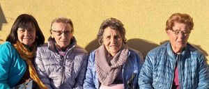 Vier ältere Frauen sitzen auf einer Bank, alle tragen Jacken und Brillen. Die Frau in der Mitte benutzt einen Stock, während eine andere einen Schal um den Hals hat. Sie lächeln und posieren für ein Foto.