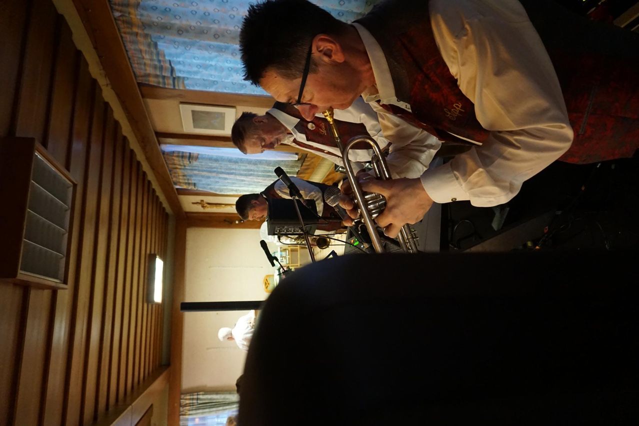 Zwei Männer spielen Trompeten in einem Raum mit Holzwänden und blauen Vorhängen. Einer trägt eine Brille und der andere eine Weste.