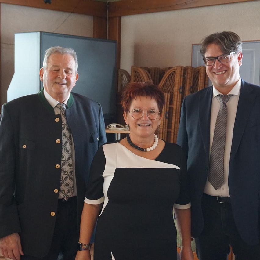 Drei Menschen stehen in einem Raum und lächeln für ein Foto. Ein Mann trägt einen blauen Anzug und eine Krawatte, ein anderer Mann trägt eine grüne Jacke und Krawatte. Eine Frau trägt ein schwarzes und weißes Kleid und eine Halskette.