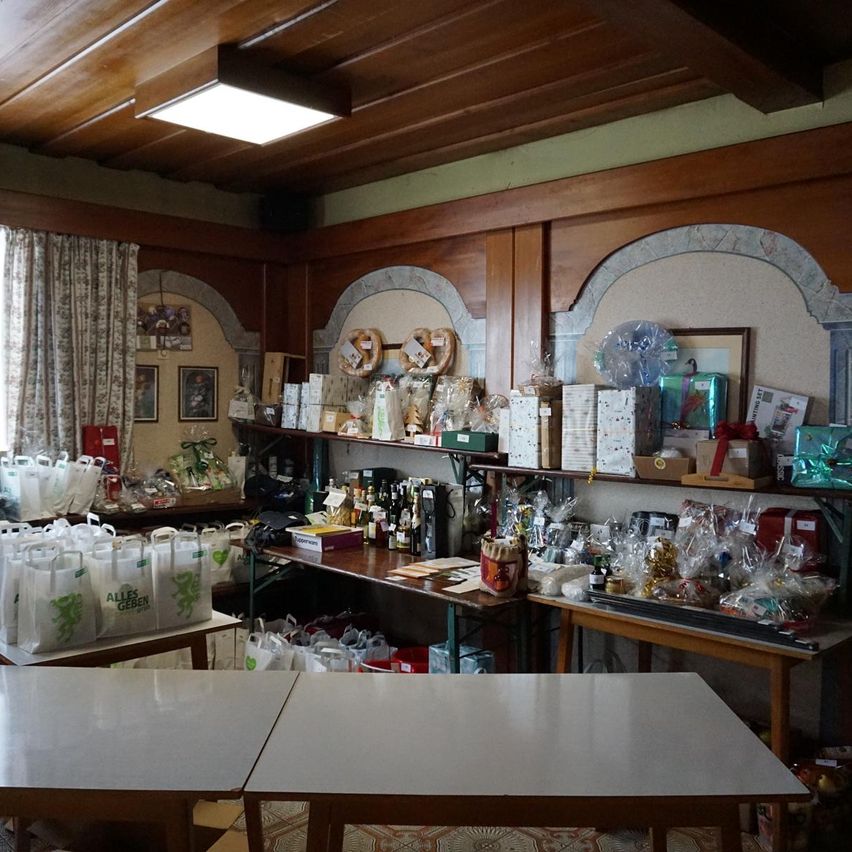 Ein Raum mit Holzwänden und Regalen, die mit verschiedenen Artikeln gefüllt sind. Die Regale sind mit zahlreichen Geschenktüten, Kisten und dekorativen Gegenständen bestückt. Ein Tisch ist mit Plastiktüten und Geschenkschachteln bedeckt. Ein weiterer Tisch im Raum ist mit Flaschen gefüllt, und es gibt eine Deckenleuchte.