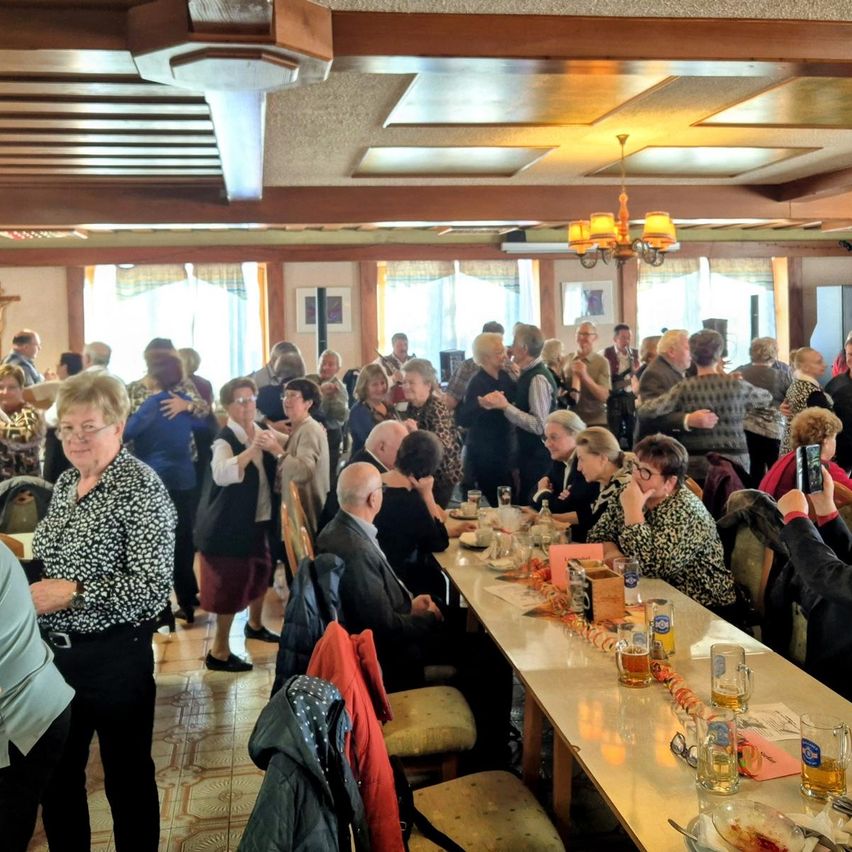 Eine Gruppe älterer Menschen versammelt sich in einem Essbereich, einige sitzen und andere stehen, mit einem Tisch voller Gläser und Essen, einer Deckenleuchte und Bilderrahmen an der Wand.