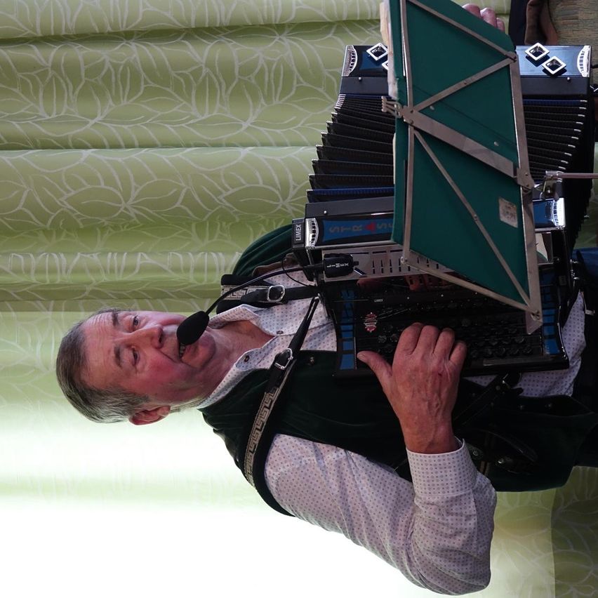 Ein älterer Mann spielt Akkordeon und benutzt ein Mikrofon im Mund, während er vor einem grünen Blumenvorhang steht.