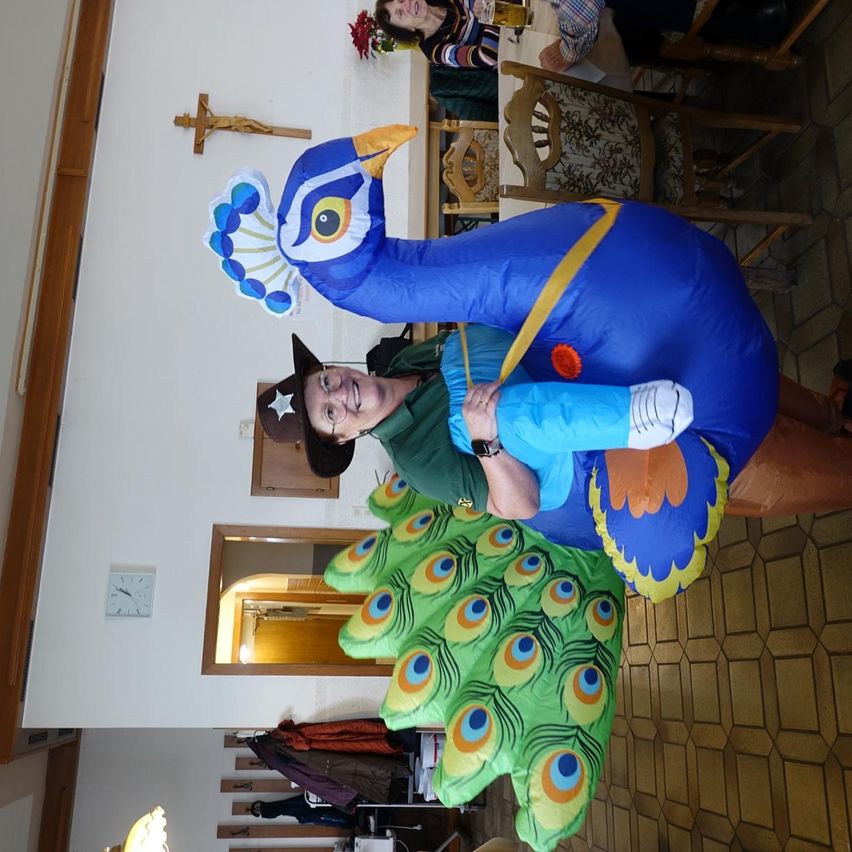 Eine Person in einem Pfauenkostüm hält ein Kind in einem blauen Kostüm. Dahinter befindet sich ein Raum mit einer weißen Wand, einem Kreuz und einem Tisch mit sitzenden Personen.