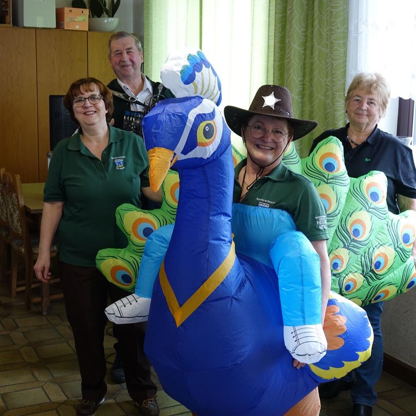 Eine Gruppe von Menschen posiert mit einem großen aufblasbaren Pfauen-Kostüm, während zwei Frauen in grünen Shirts und eine Frau in einer Cowboy-Mütze lächeln. Ein Mann steht hinter ihnen in der Nähe eines Holzschranks mit einer Pflanze.
