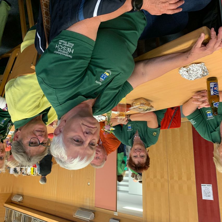 Mehrere Personen in grünen Shirts sitzen an einem Tisch, einer mit einer Bierflasche und anderen lächelnd. Eine Person hat ein Getränk und ein Sandwich.