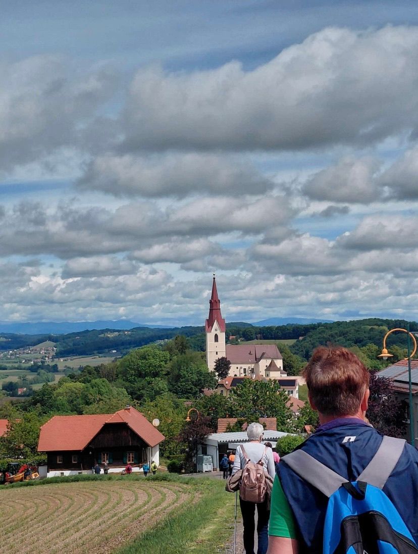 Die Aussicht von einem Hügel zeigt eine Stadt mit einem Kirchturm, Häusern und Bäumen unter einem bewölkten Himmel. Menschen gehen durch die Stadt, und ein Mann betrachtet die Aussicht.