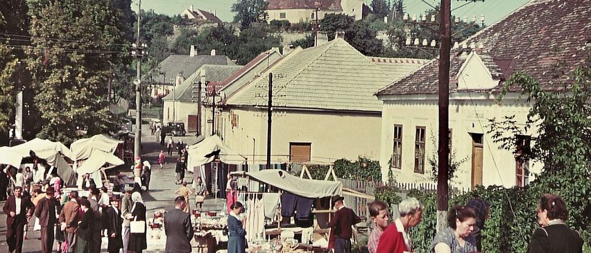 Ein altes Foto zeigt eine belebte Marktstraße mit einkaufenden Menschen. Es gibt Zelte, Stände und verschiedene zu verkaufende Artikel. Traditionelle Gebäude säumen die Straße.