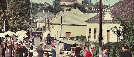 Ein Vintage-Foto eines geschäftigen Straßenmarktes mit einkaufenden Menschen. Gebäude säumen die Straße, einige mit Vordächern. Einige Personen betrachten ausgestellte Artikel, während andere in Gespräche vertieft sind.