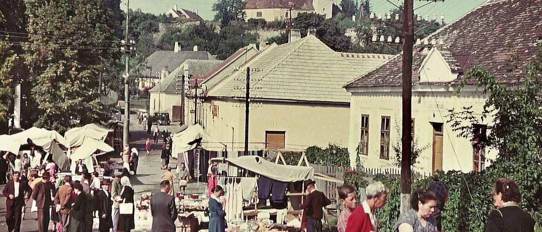 Ein Vintage-Foto eines geschäftigen Straßenmarktes mit einkaufenden Menschen. Gebäude säumen die Straße, einige mit Vordächern. Einige Personen betrachten ausgestellte Artikel, während andere in Gespräche vertieft sind.