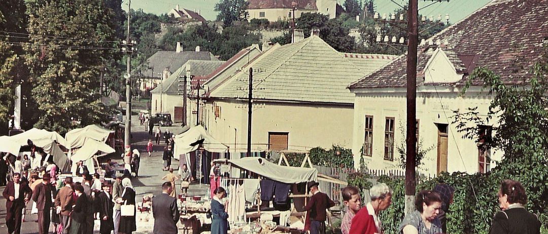 Ein Vintage-Foto eines geschäftigen Straßenmarktes mit einkaufenden Menschen. Gebäude säumen die Straße, einige mit Vordächern. Einige Personen betrachten ausgestellte Artikel, während andere in Gespräche vertieft sind.