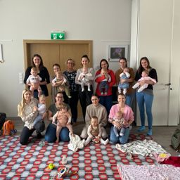 Eine Gruppe lächelnder Mütter und ihrer Säuglinge in einem Raum, wobei einige sitzen und andere stehen. Einige Babys werden gehalten und ein Teppich mit Spielzeug liegt auf dem Boden.