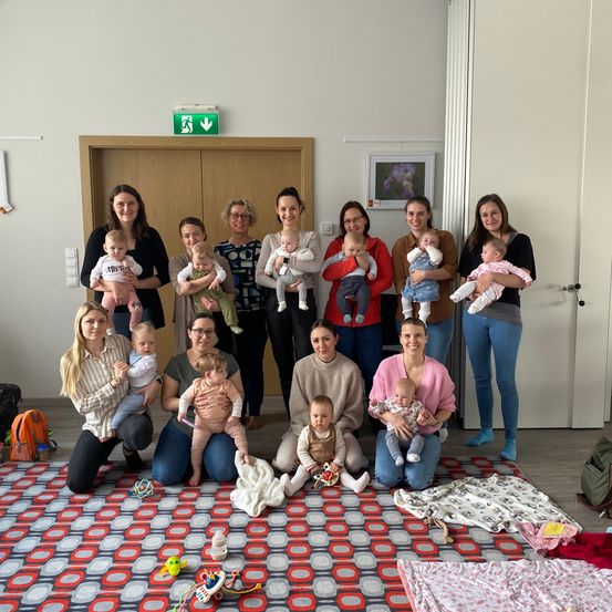 Eine Gruppe lächelnder Mütter und ihrer Säuglinge in einem Raum, wobei einige sitzen und andere stehen. Einige Babys werden gehalten und ein Teppich mit Spielzeug liegt auf dem Boden.