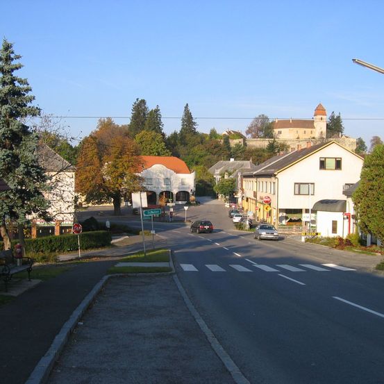 Ein kleines Dorf mit einer Burg in der Ferne. Die Straße ist leer, mit geparkten Autos an der Seite. Bäume und Büsche säumen die Straße.