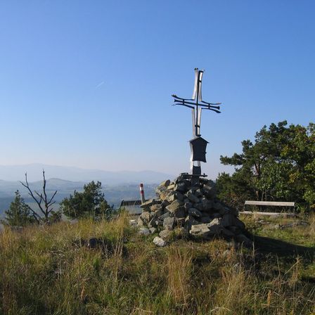 Ein Kreuz auf einem Steinhaufen auf einem grasigen Hügel, mit einer Bank und Bäumen im Hintergrund unter einem klaren blauen Himmel.