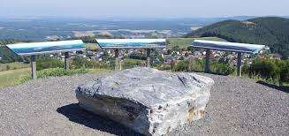 Drei Schilder stehen auf einem felsigen Hügel und bieten einen Panoramablick auf ein Tal mit einer kleinen Stadt darunter. Ein großer Felsblock liegt im Vordergrund.