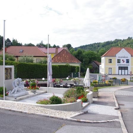 Eine Straßenszene mit einer Gedenkskulptur im Vordergrund, umgeben von Blumen. Dahinter ist eine Feuerwehr mit einem Schild 'Feuerwehr Gemeindehaus' zu sehen. In der Nähe befinden sich Häuser mit roten Dächern und ein Garten. Der Himmel ist bewölkt.