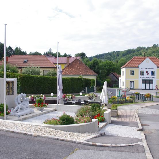 Eine Straßenszene mit einer Gedenkskulptur im Vordergrund, umgeben von Blumen. Dahinter ist eine Feuerwehr mit einem Schild 'Feuerwehr Gemeindehaus' zu sehen. In der Nähe befinden sich Häuser mit roten Dächern und ein Garten. Der Himmel ist bewölkt.