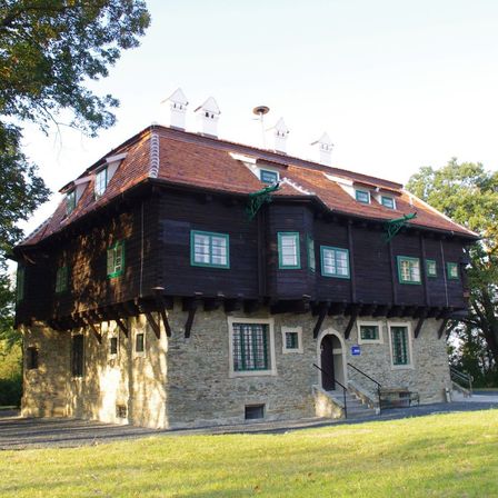 Ein großes Holzgebäude mit braunem Dach, grünen Fenstern und Backsteinwänden. Es hat mehrere Ebenen und Schornsteine. Das Gebäude ist von einem grünen Rasen und Bäumen umgeben.