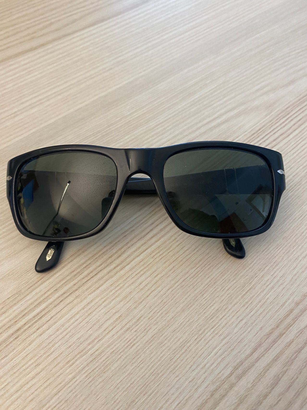 Ein Paar schwarze Sonnenbrille mit rechteckigen Gläsern und einem dünnen schwarzen Rahmen liegt auf einer Holzoberfläche.