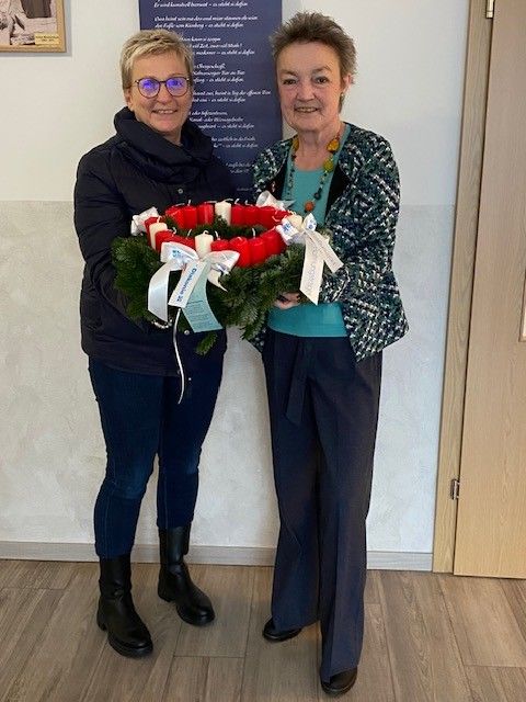 Zwei Frauen stehen nebeneinander und halten einen Kranz mit roten und weißen Kerzen und einem Band. Sie lächeln und schauen nach vorne. Hinter ihnen befindet sich ein Poster mit fremdsprachigem Text und eine Tür.